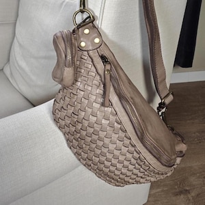 Puede incluir: Bolso bandolera de cuero tejido en color topo neutro. El bolso tiene un compartimento principal con cierre de cremallera, un bolsillo peque&ntilde;o adicional con cremallera y una correa de cuero marr&oacute;n. El bolso est&aacute; colgado del brazo de una silla blanca. Mide unos 30 cm de largo.