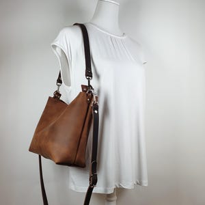 Op de afbeelding: Een bruine leren schoudertas met een lange, verstelbare riem. De tas wordt getoond op een witte mannequin, die de vorm en het ontwerp laat zien. De kleur van de tas is een rijk, warm bruin, en de riem is een donkerdere tint.