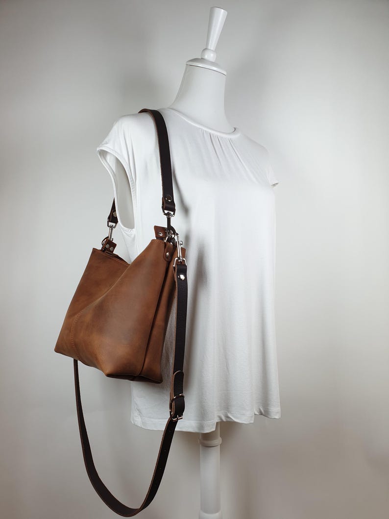 Op de afbeelding: Een bruine leren schoudertas met een lange, verstelbare riem. De tas wordt getoond op een witte mannequin, die de vorm en het ontwerp laat zien. De kleur van de tas is een rijk, warm bruin, en de riem is een donkerdere tint.