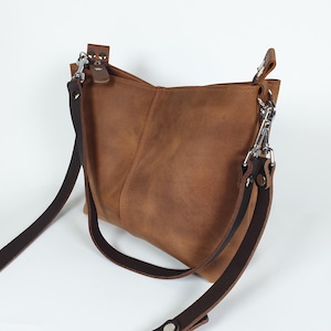 Op de afbeelding: Een bruine leren crossbody tas met een verstelbare donkerbruine riem en zilverkleurige hardware. De tas heeft een rechthoekige vorm met een licht afgeronde bodem en een klepsluiting. Het leer is zacht en getextureerd.