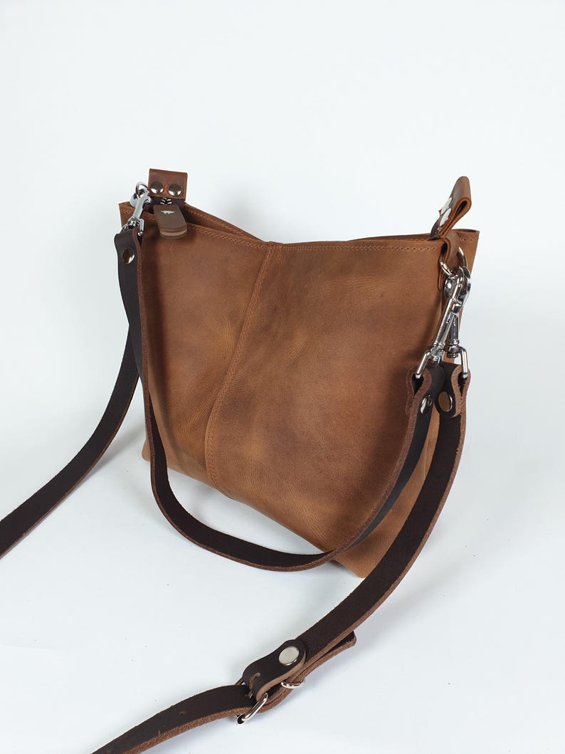 Op de afbeelding: Een bruine leren crossbody tas met een verstelbare donkerbruine riem en zilverkleurige hardware. De tas heeft een rechthoekige vorm met een licht afgeronde bodem en een klepsluiting. Het leer is zacht en getextureerd.