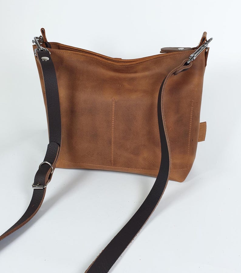 Op de afbeelding: Bruine leren schoudertas met een verstelbare donkerbruine riem en zilverkleurige hardware. De tas heeft een rechthoekige vorm met een licht gebogen bovenkant en een verticaal gestikt detail aan de voorkant.