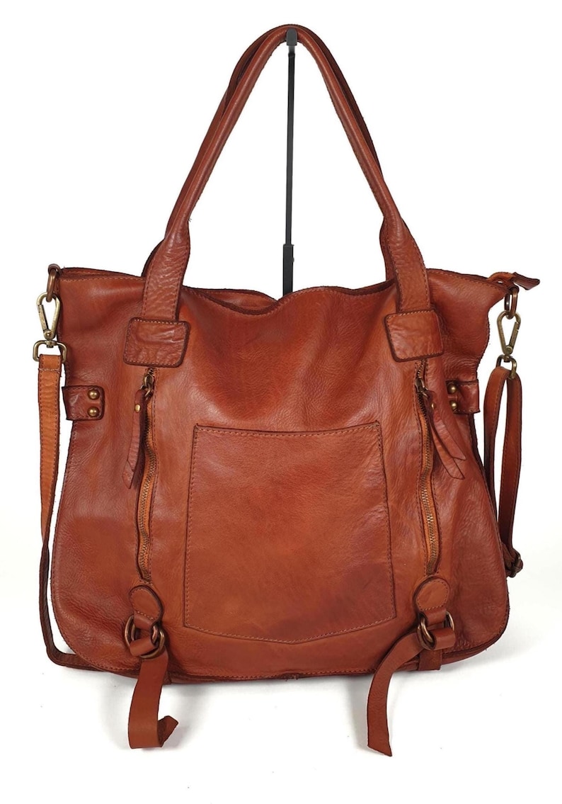 May include: A brown leather tote bag with two top handles and a detachable shoulder strap. The bag has a front pocket, two zippered compartments, and metal hardware. The leather has a distressed look.