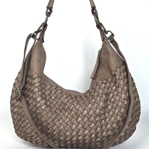 May include: A woven, taupe-colored hobo bag with a shoulder strap and a crossbody strap. The bag features a large, curved design with a basket-weave pattern. The straps are attached with metal rings.