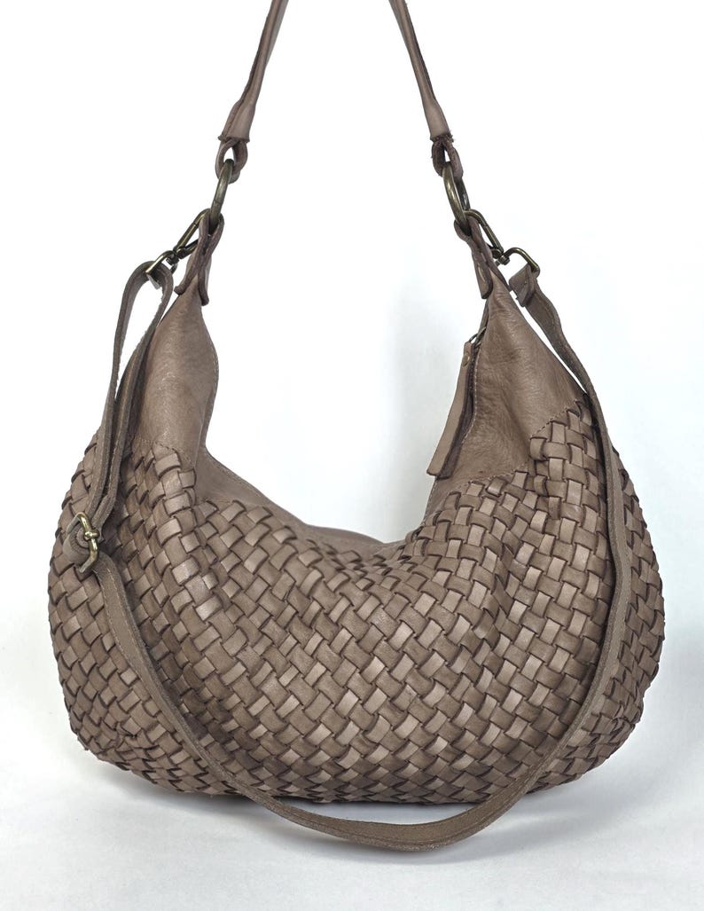 May include: A woven, taupe-colored hobo bag with a shoulder strap and a crossbody strap. The bag features a large, curved design with a basket-weave pattern. The straps are attached with metal rings.