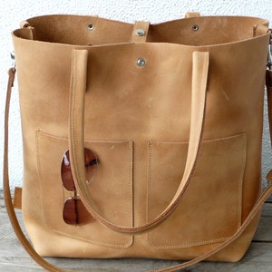 Puede incluir: Un bolso tote grande de cuero marr&oacute;n claro con dos asas superiores y una correa larga para el hombro. El bolso tiene dos bolsillos grandes en la parte delantera y un par de gafas de sol est&aacute;n metidas en uno de los bolsillos.