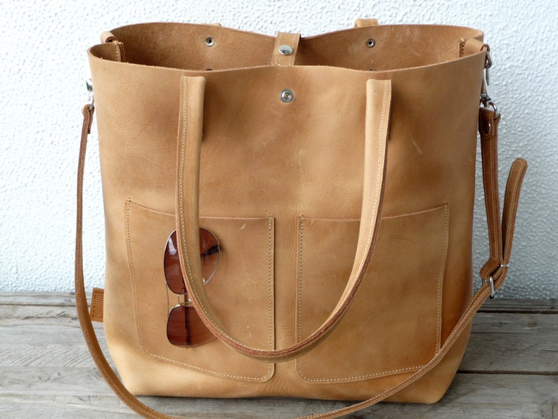 Puede incluir: Un bolso tote grande de cuero marr&oacute;n claro con dos asas superiores y una correa larga para el hombro. El bolso tiene dos bolsillos grandes en la parte delantera y un par de gafas de sol est&aacute;n metidas en uno de los bolsillos.