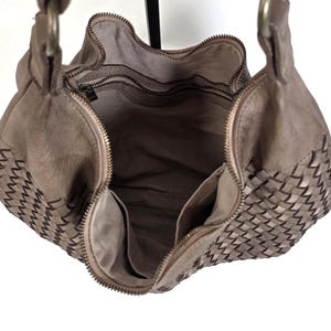 May include: Taupe leather hobo bag with woven detailing on the sides. The bag features a zipper closure and two interior pockets. The handles are attached with metal rings. The bag is open, revealing the interior lining.