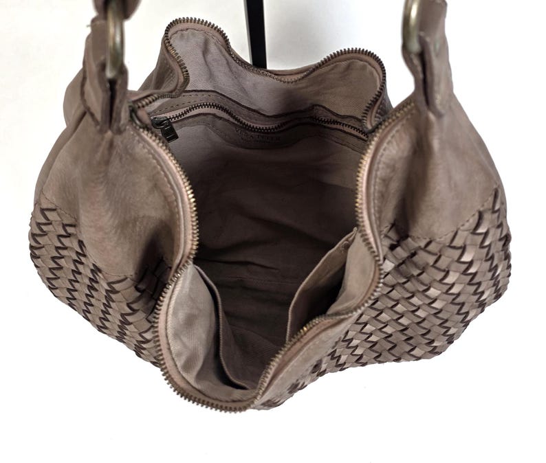 May include: Taupe leather hobo bag with woven detailing on the sides. The bag features a zipper closure and two interior pockets. The handles are attached with metal rings. The bag is open, revealing the interior lining.