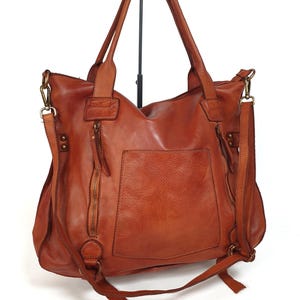 May include: A brown leather tote bag with a shoulder strap and a front pocket. The bag has a zipper closure and a top handle. The leather has a distressed look. The bag is displayed on a black stand against a white background.