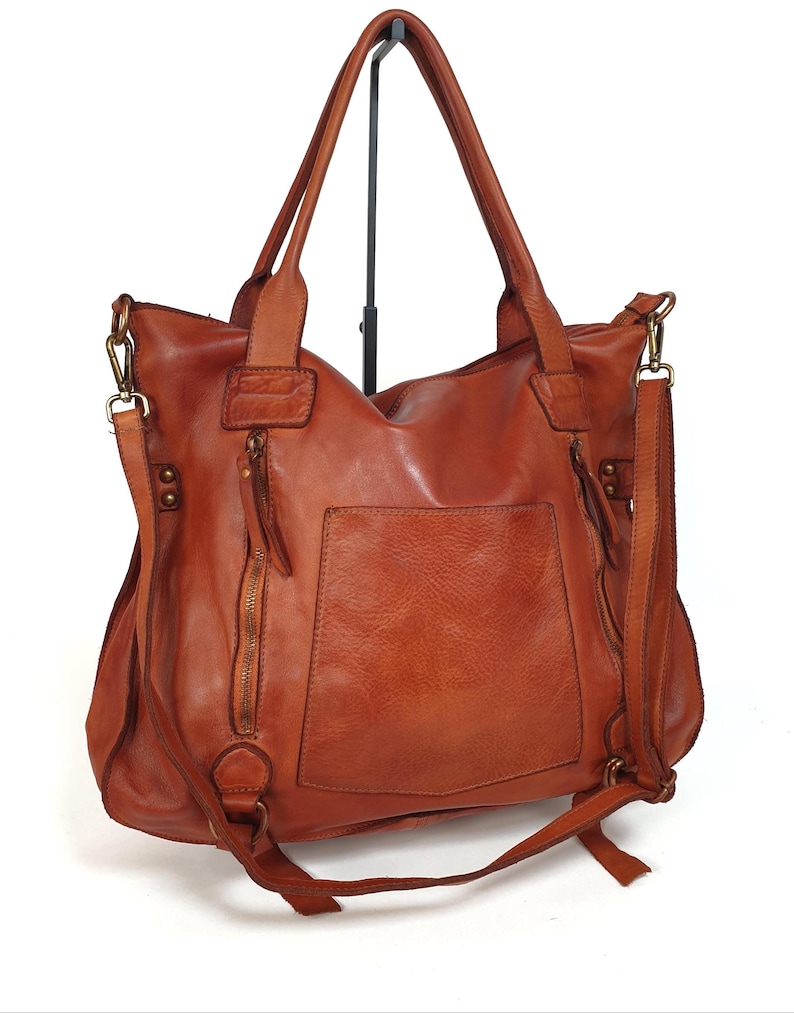May include: A brown leather tote bag with a shoulder strap and a front pocket. The bag has a zipper closure and a top handle. The leather has a distressed look. The bag is displayed on a black stand against a white background.