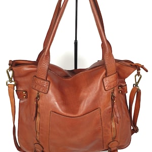 May include: A brown leather tote bag with two top handles and a detachable shoulder strap. The bag features two zippered pockets on the front and a large pocket on the back. The bag is made of a distressed leather.