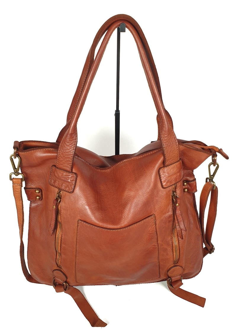 May include: A brown leather tote bag with two top handles and a detachable shoulder strap. The bag features two zippered pockets on the front and a large pocket on the back. The bag is made of a distressed leather.