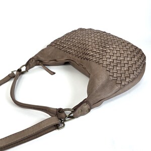 May include: A taupe-colored shoulder bag with a woven design on the top section. The bag has a long, adjustable strap and a zipper closure. The hardware is a dark, antique metal. The bag is made of a soft, leather-like material.