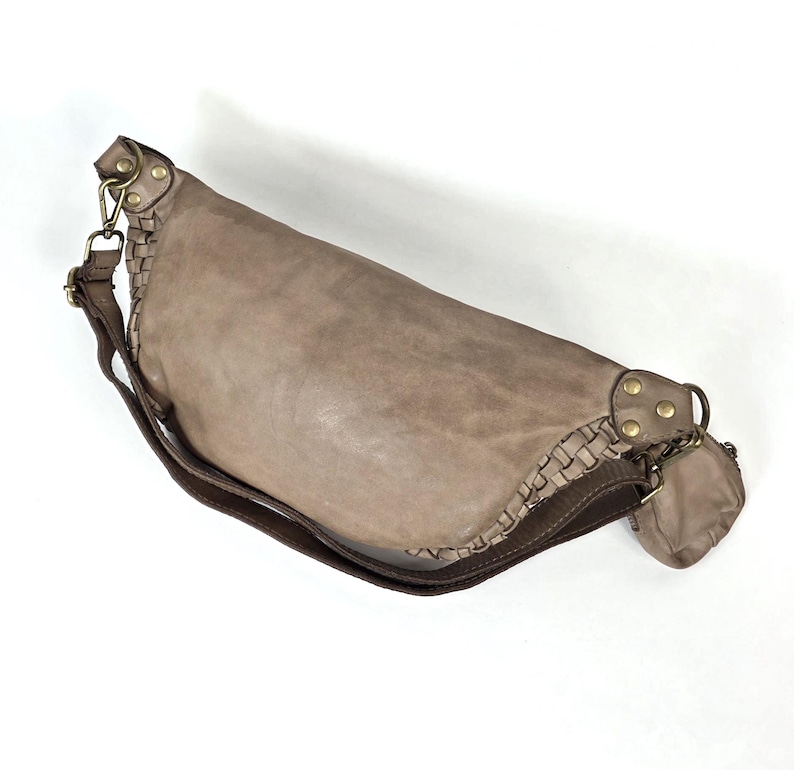 Puede incluir: Bolso de hombro de cuero color topo con un detalle trenzado en el lateral. Tiene una correa larga y ajustable y una peque&ntilde;a bolsa adjunta. Los herrajes son de color lat&oacute;n.
