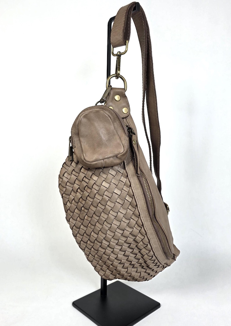 Puede incluir: Bolso bandolera con estampado tejido en color topo neutro. El bolso presenta un peque&ntilde;o bolsillo adjunto y una correa larga y ajustable. El dise&ntilde;o incluye cierre de cremallera y herrajes met&aacute;licos. El bolso se exhibe en un soporte negro.