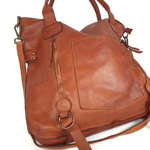May include: A brown leather handbag with a shoulder strap and top handles. The bag has a large front pocket, a zipper closure, and brass-colored hardware. The leather has a distressed appearance.