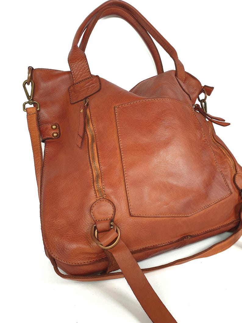 May include: A brown leather handbag with a shoulder strap and top handles. The bag has a large front pocket, a zipper closure, and brass-colored hardware. The leather has a distressed appearance.