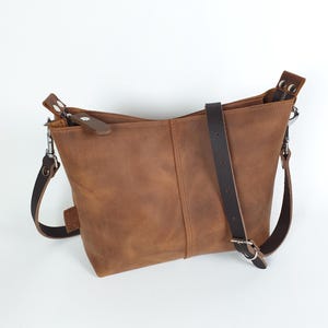 Op de afbeelding: Bruine leren crossbodytas met een verstelbare donkerbruine riem en zilverkleurige hardware. De tas heeft een rechthoekige vorm met een ritssluiting en een klein leren accent op de ritstrekker. De tas is gemaakt van leer.