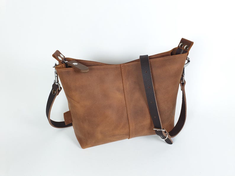 Op de afbeelding: Bruine leren crossbodytas met een verstelbare donkerbruine riem en zilverkleurige hardware. De tas heeft een rechthoekige vorm met een ritssluiting en een klein leren accent op de ritstrekker. De tas is gemaakt van leer.