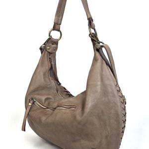 May include: A taupe-colored leather hobo bag with a single shoulder strap. The bag features a zipper closure, a front pocket with a zipper, and decorative stitching along the side. The bag has brass-colored metal hardware.