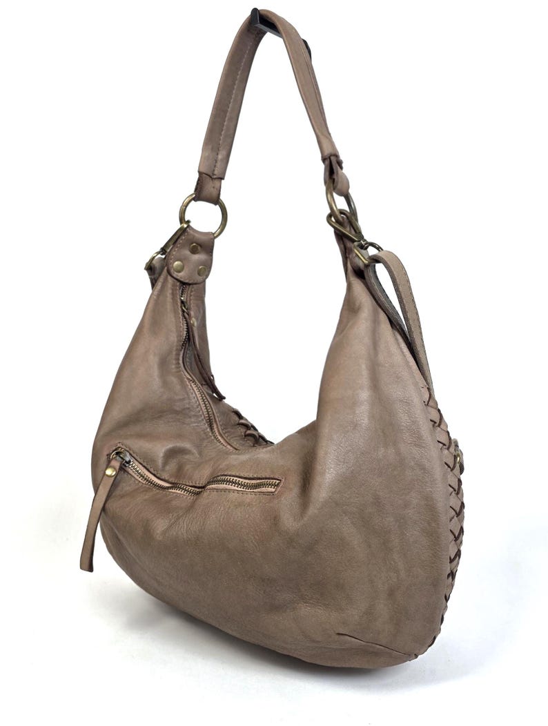 May include: A taupe-colored leather hobo bag with a single shoulder strap. The bag features a zipper closure, a front pocket with a zipper, and decorative stitching along the side. The bag has brass-colored metal hardware.