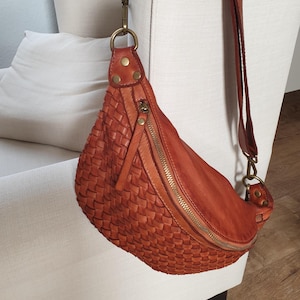 Pode incluir: Uma bolsa transversal de couro tecido em um tom marrom avermelhado quente. A bolsa tem formato curvo, uma longa alça ajustável e fecho de zíper. A bolsa está pendurada em uma cadeira branca.