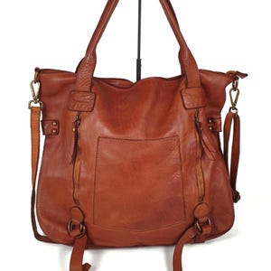 Das Bild zeigt eine große Leder Tragetasche in cognac braun der Marke sanumileathergoods. Diese Tasche ist 44 cm breit, 37 cm hoch und 13 cm tief und hängt mit beiden Schulterhenkeln an einem Taschenständer vor weißem Hintergrund. Diese weiche Tote Bag schließt oben mit einem Reißverschluss und hat zusätzlich zwei kleine Reißverschlusstaschen senkrecht auf der Vorderseite. In der Mitte dazwischen befindet sich ein kleines offenes Eingrifffach. Zusätzlich hat die Tasche einen langen Umhängegurt.