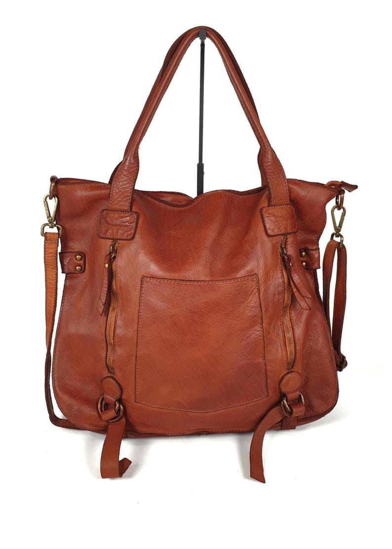 Das Bild zeigt eine große Leder Tragetasche in cognac braun der Marke sanumileathergoods. Diese Tasche ist 44 cm breit, 37 cm hoch und 13 cm tief und hängt mit beiden Schulterhenkeln an einem Taschenständer vor weißem Hintergrund. Diese weiche Tote Bag schließt oben mit einem Reißverschluss und hat zusätzlich zwei kleine Reißverschlusstaschen senkrecht auf der Vorderseite. In der Mitte dazwischen befindet sich ein kleines offenes Eingrifffach. Zusätzlich hat die Tasche einen langen Umhängegurt.