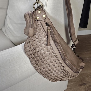 Pode incluir: Bolsa transversal de couro trançado marrom e bege. Possui um compartimento principal com zíper, uma alça ajustável e um pequeno bolso com zíper. A bolsa está pendurada em uma cadeira branca.