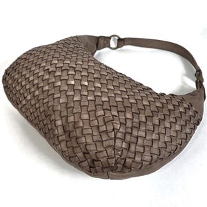 May include: A woven, taupe-colored shoulder bag with a curved shape. The bag features a braided design and a matching strap with a metal ring attachment. The bag is made of a soft material and is suitable for everyday use.
