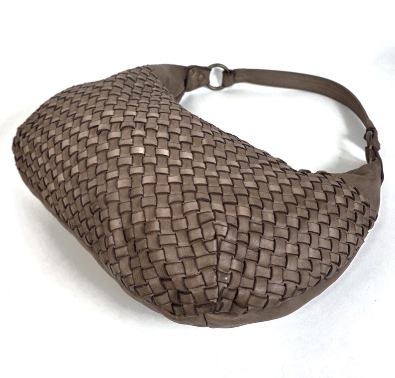 May include: A woven, taupe-colored shoulder bag with a curved shape. The bag features a braided design and a matching strap with a metal ring attachment. The bag is made of a soft material and is suitable for everyday use.