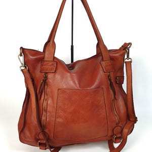 May include: A brown leather tote bag with a front pocket and two zippered compartments. The bag has two top handles and a detachable shoulder strap. The bag is displayed on a black stand against a white background.
