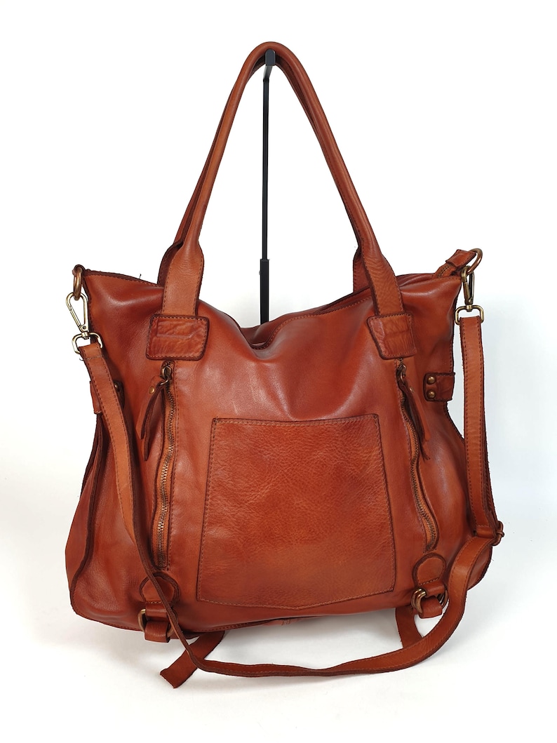 May include: A brown leather tote bag with a front pocket and two zippered compartments. The bag has two top handles and a detachable shoulder strap. The bag is displayed on a black stand against a white background.