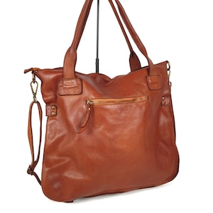 May include: A brown leather tote bag with two top handles and a detachable shoulder strap. The bag features a zippered closure on the back and a front pocket. The leather has a distressed look, and the bag appears to be new.