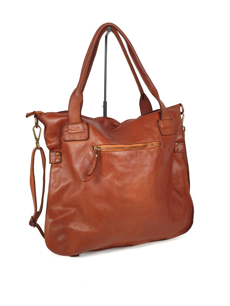 May include: A brown leather tote bag with two top handles and a detachable shoulder strap. The bag features a zippered closure on the back and a front pocket. The leather has a distressed look, and the bag appears to be new.