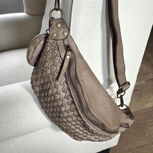 Puede incluir: Ri&ntilde;onera de cuero tejido en color topo neutro. La bolsa tiene un compartimento principal con cremallera, un peque&ntilde;o bolsillo con cremallera y una correa ajustable. La bolsa est&aacute; colgada en una silla blanca.