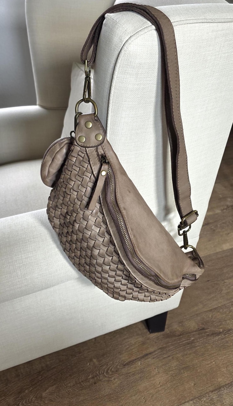 Puede incluir: Ri&ntilde;onera de cuero tejido en color topo neutro. La bolsa tiene un compartimento principal con cremallera, un peque&ntilde;o bolsillo con cremallera y una correa ajustable. La bolsa est&aacute; colgada en una silla blanca.