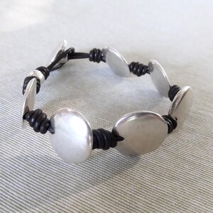 Puede incluir: Un brazalete de plata con abalorios redondos de metal unidos por un cord&oacute;n de cuero negro.