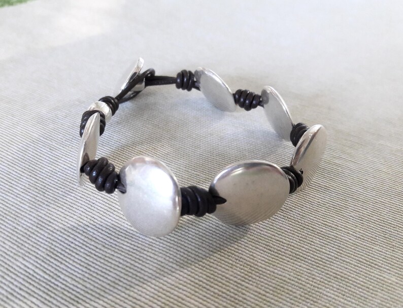 Puede incluir: Un brazalete de plata con abalorios redondos de metal unidos por un cord&oacute;n de cuero negro.