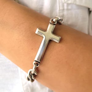 Puede incluir: Un brazalete de cadena de plata con un gran colgante de cruz de plata.