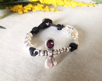 May include: A black leather bracelet with silver beads and a silver charm featuring a purple gemstone. The charm is shaped like a cross.