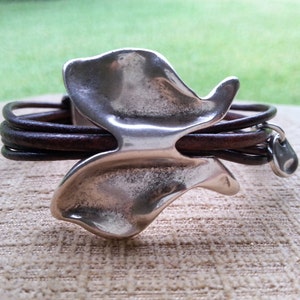 May include: A brown leather bracelet with a silver butterfly charm. The butterfly charm has a textured, organic shape.