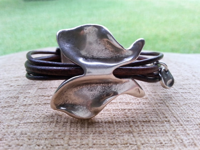 May include: A brown leather bracelet with a silver butterfly charm. The butterfly charm has a textured, organic shape.