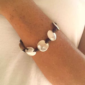 Puede incluir: Un brazalete de cuero marr&oacute;n con abalorios redondos plateados. El brazalete est&aacute; en la mu&ntilde;eca de una persona.