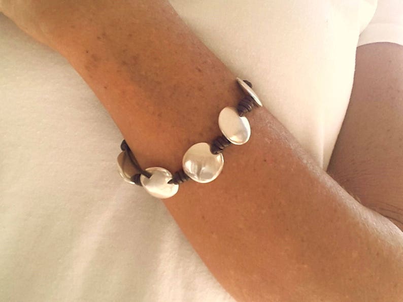 Puede incluir: Un brazalete de cuero marr&oacute;n con abalorios redondos plateados. El brazalete est&aacute; en la mu&ntilde;eca de una persona.