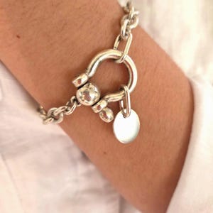 May include: A silver-toned bracelet featuring a large U-shaped clasp and a small, round charm. The bracelet has a chunky chain design and is worn on a person's wrist, against a white shirt.