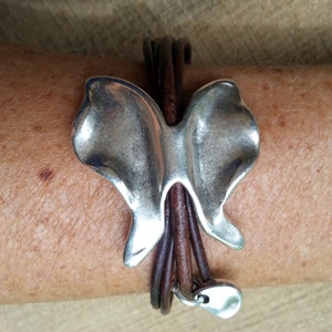 May include: A silver butterfly charm bracelet with brown leather straps. The bracelet has a silver heart charm at the end.