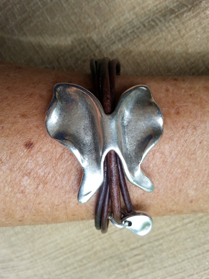 May include: A silver butterfly charm bracelet with brown leather straps. The bracelet has a silver heart charm at the end.