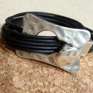 May include: A black leather bracelet with a silver, textured, abstract-shaped clasp.